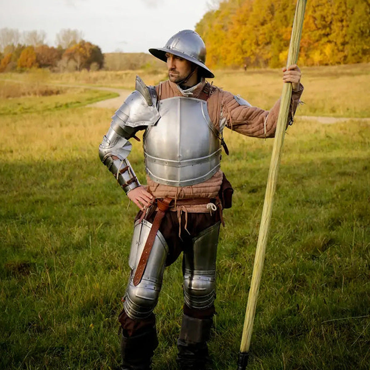 A knight in armor equipped with padded protectors, standing confidently in a green field with autumn trees.