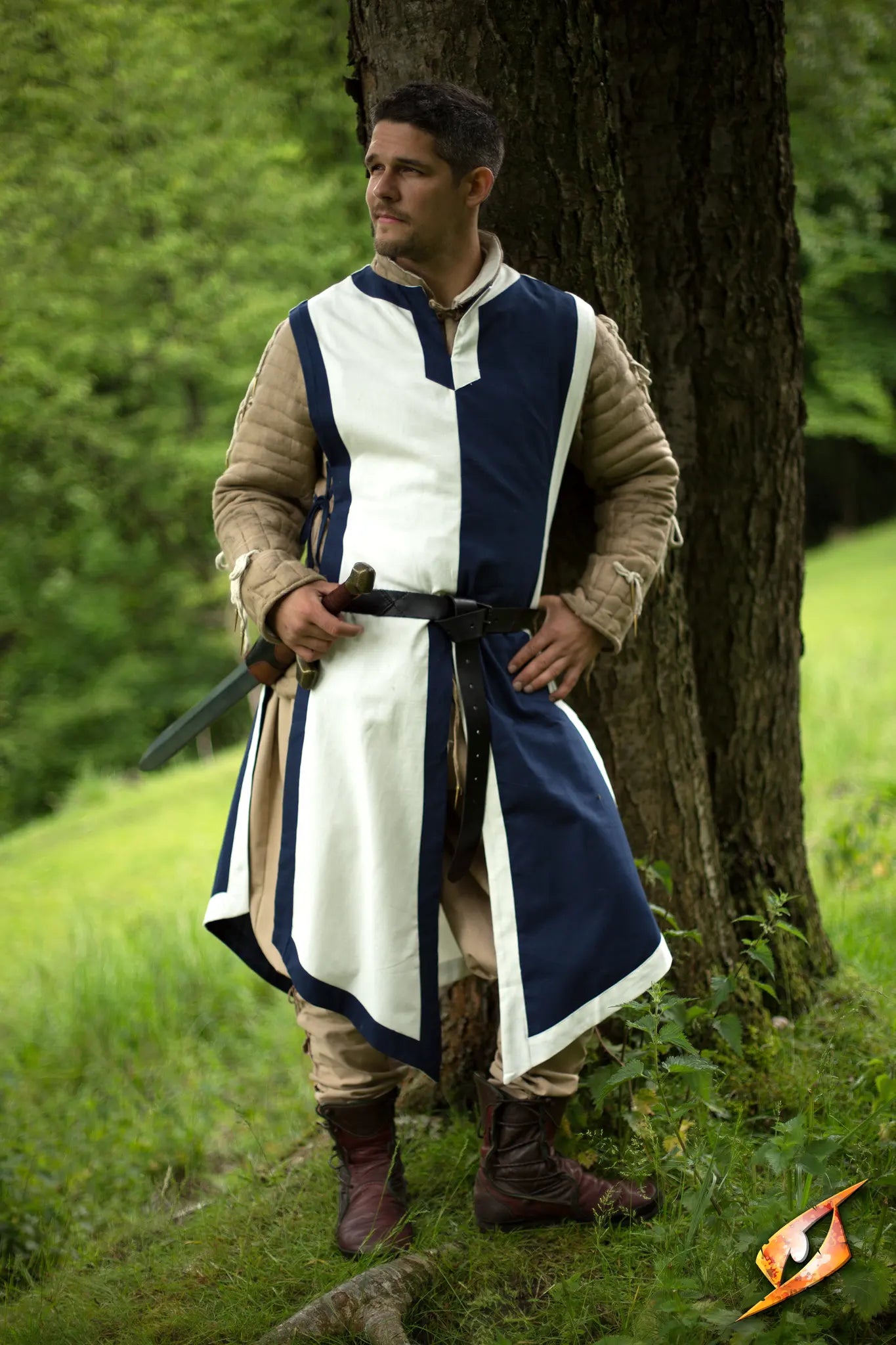 A man wearing a Basic Tabard stands confidently by a tree, ready for action in a medieval setting.