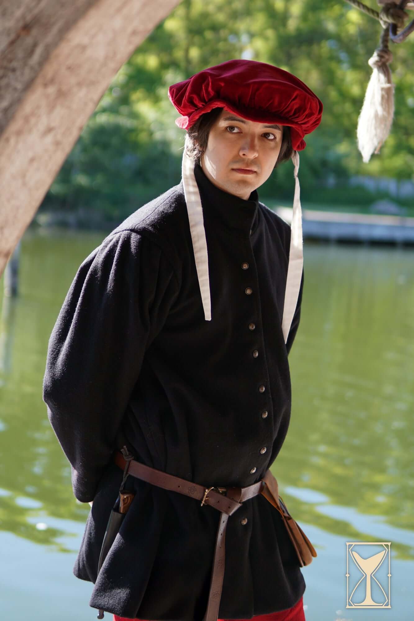 Surcoat Lorenzo worn by a male model, featuring traditional 14th century design and style, set by a tranquil water backdrop.