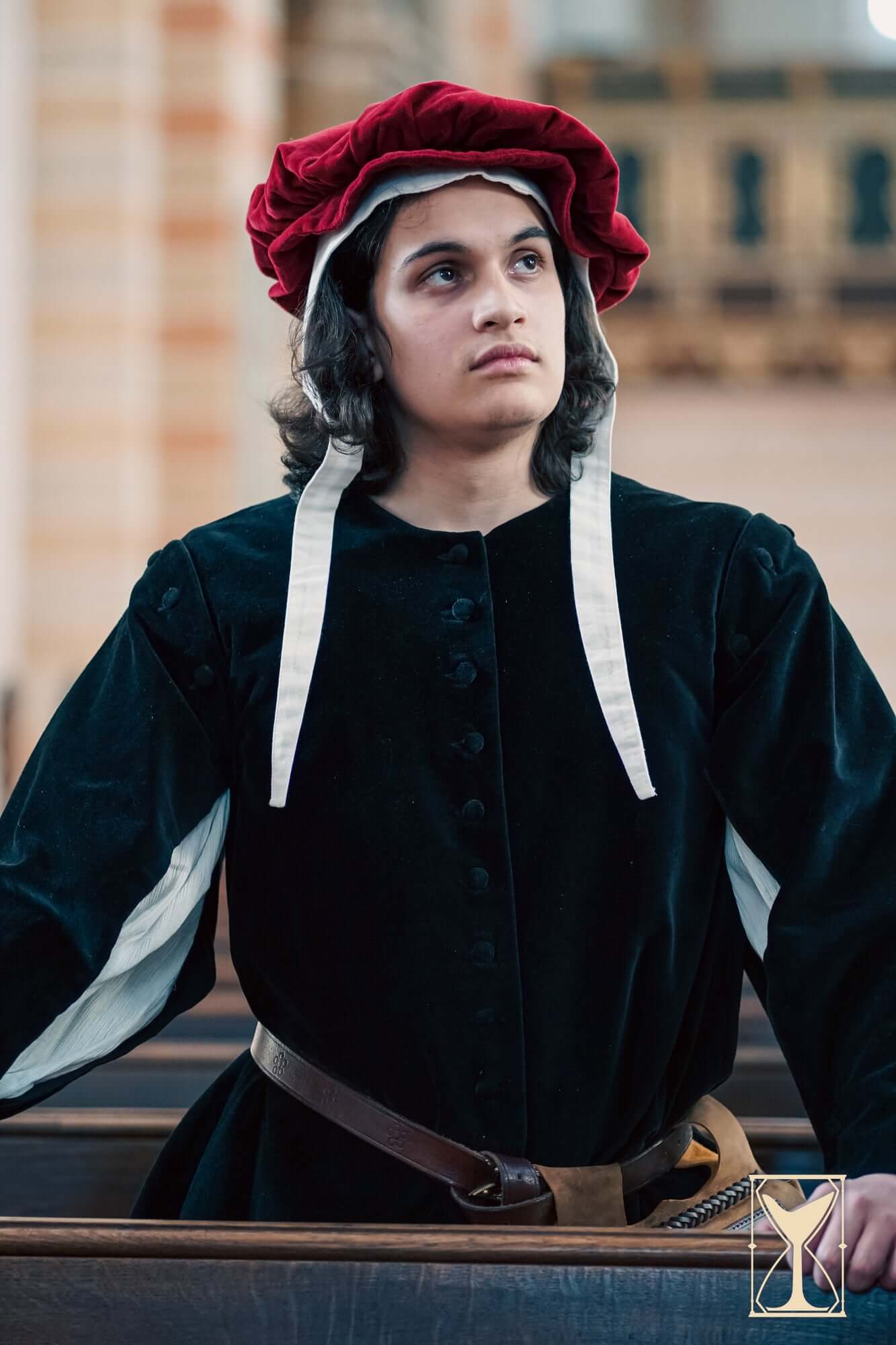 Young man in a velvet surcoat and red hat, embodying medieval fashion in a church setting.