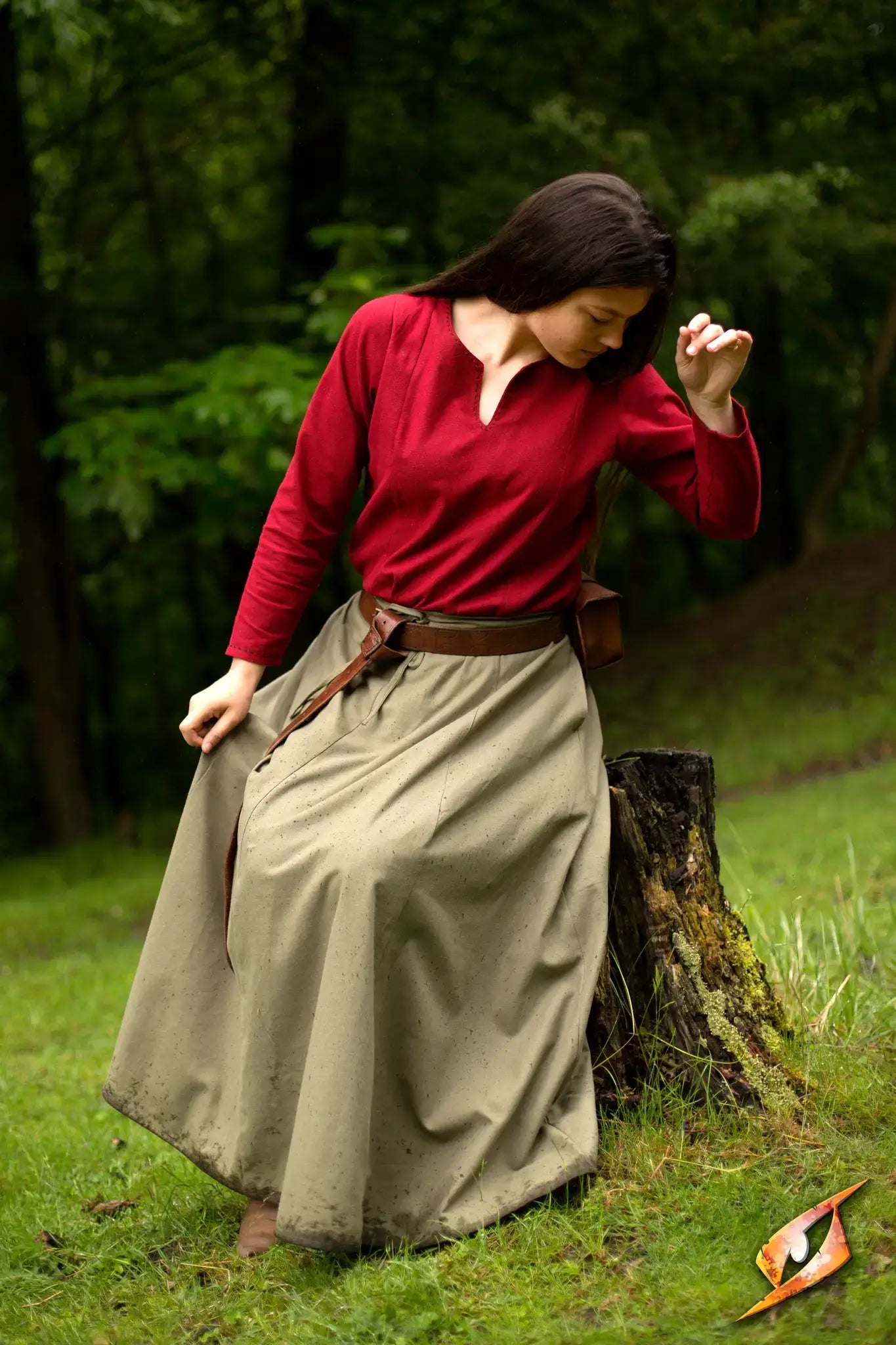 A woman wearing a red top and a flowing medieval skirt sitting on a stump outdoors.