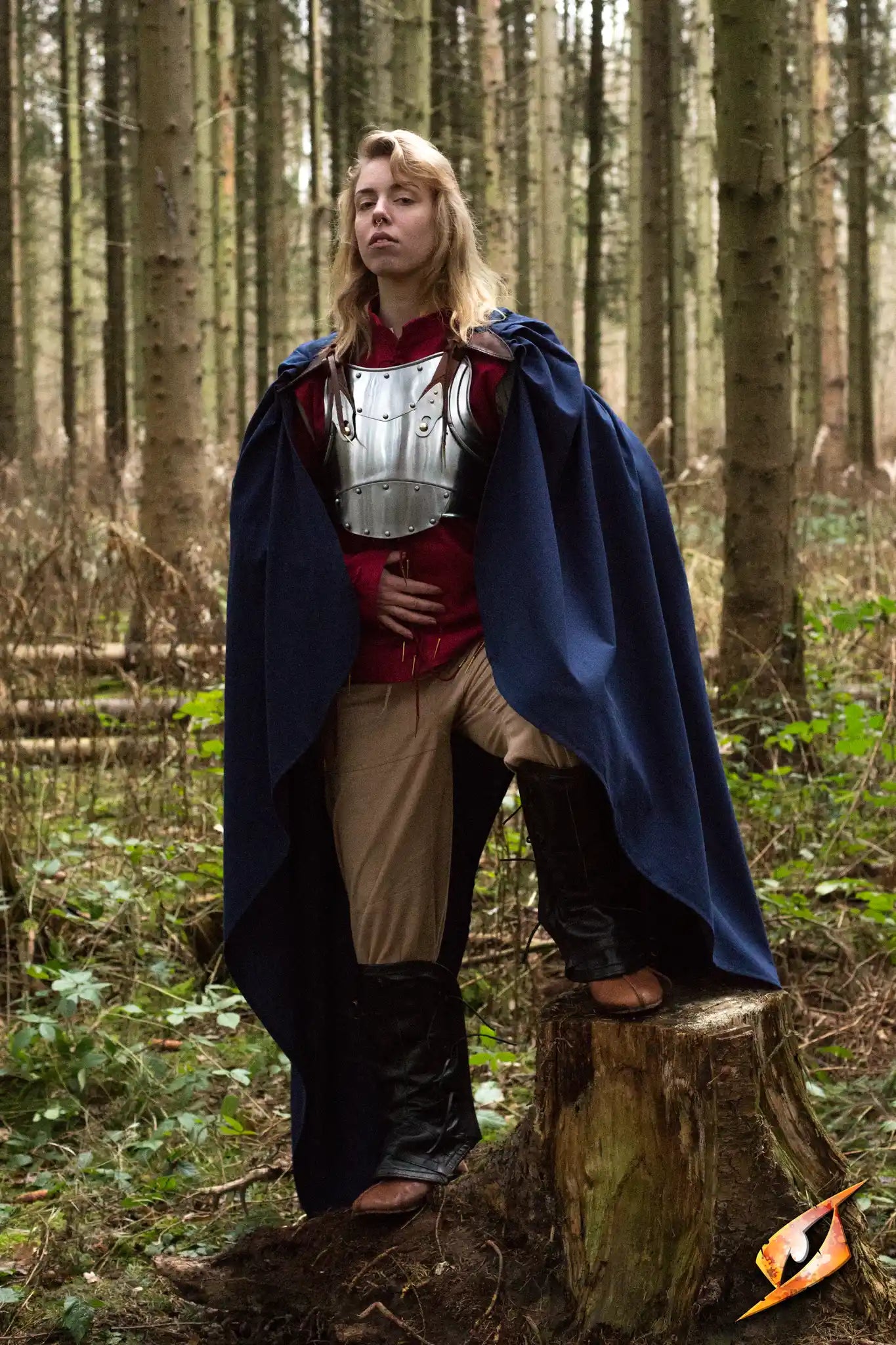 A model wearing the Cape Regulus, a medium-length blue cape, in a forest setting, showcasing its elegant design and protection.