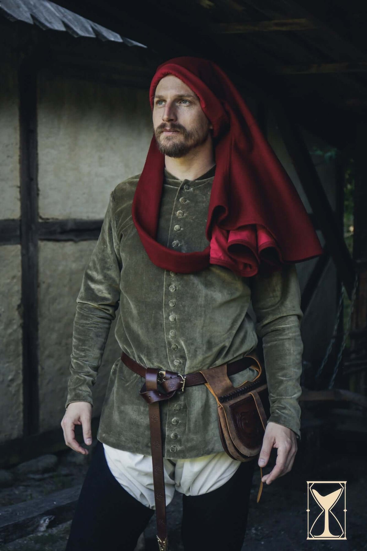 Cotehardie Bonifacio, a man wears a green cotehardie with a red hood and a leather belt, set in a rustic environment.